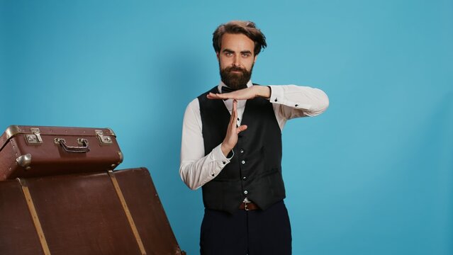 Hotel concierge does timeout symbol in studio, asking for a break working in hotel and tourism industry. Elegant doorkeeper in formal attire indicating pause gesture with hands, professional job.