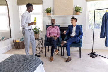 Obraz premium Three male friends chatting in bedroom dressing room, holding bottle and glass near suit rack