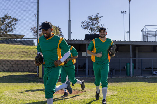 Three male baseball teammates running across grass field wearing green yellow uniforms and gloves