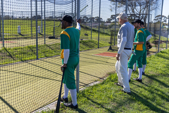 Coach signaling start, batter leaning on net holding bat, teammates watching preparing for practice