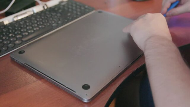 Hands of a craftsman use a blue plastic spatula tool to open the bottom cover of a laptop case