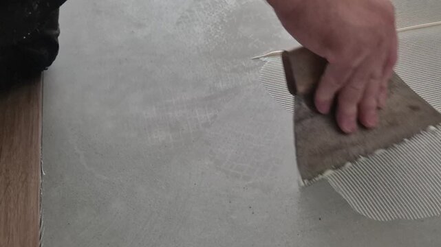 Slow motion close-up of spreading parquet adhesive on a concrete subfloor using a notched metal spatula, floor renovation process.