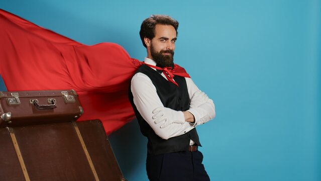 Hotel worker with red mantle to help guests with trolley bags, pretending to be fantasy cartoon superhero character on camera. Bellhop acting like comic superhuman with cape against background.