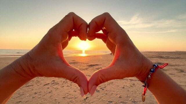 Personal perspective of a woman's hands making a heart shape, framing the beautiful sun setting over the ocean, symbolizing love, happiness, freedom, and enjoying a peaceful vacation