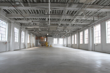 Obraz premium Large, empty, bright interior space with high ceilings, exposed beams, numerous windows, and a smooth concrete floor. The room is light