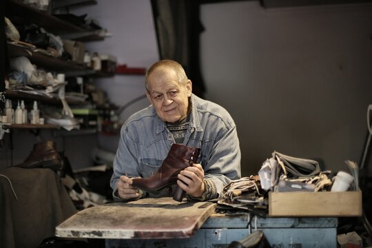 Elderly artisan holds a boot at his workbench