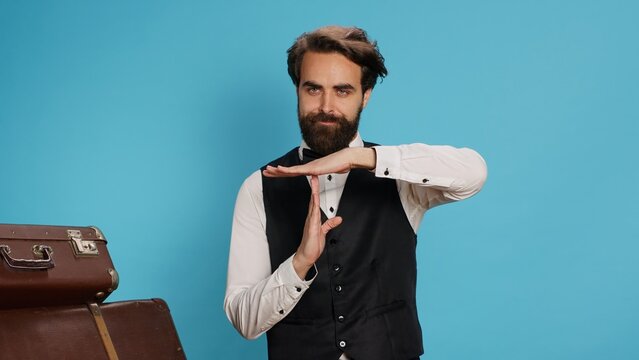 Hotel worker indicates timeout sign in studio, asking for work break and presenting hospitality and travel sector. Stylish doorkeeper in formal clothes showcasing pause symbol with hands.