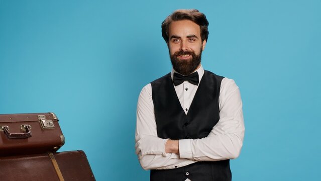 Professional hotel concierge posing on camera against blue background, preparing to carry luggage and help travellers. Elegant skilled employee working as bellboy, tourism industry concept.