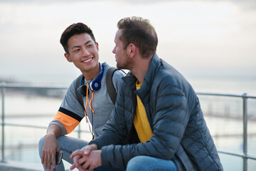Men, friends and chat on promenade at beach with smile, bonding and conversation on seaside vacation. People, relax and holiday with discussion, memory or tourism with break on getaway by ocean © peopleimages.com