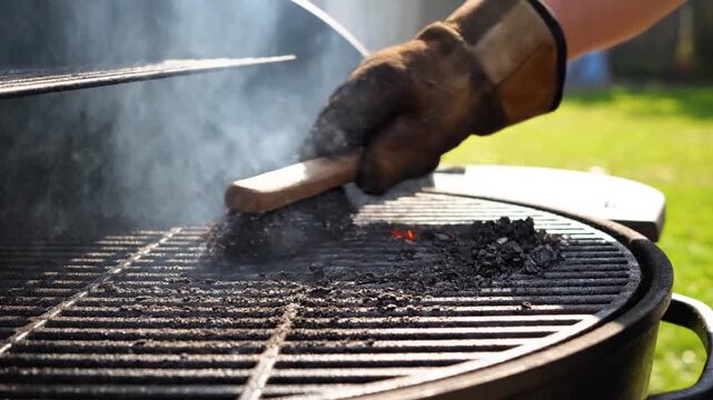 Hand in heat resistant glove cleaning a smoking charcoal grill with a wire brush.