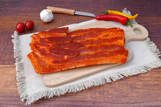 Roher eingelegter Schweinebauch auf einem Holzschneidebrett mit Stoffunterlage. Im Hintergrund liegen Knoblauch, Kirschtomaten, Chilischoten und ein K&uuml;chenmesser auf einem Holztisch.