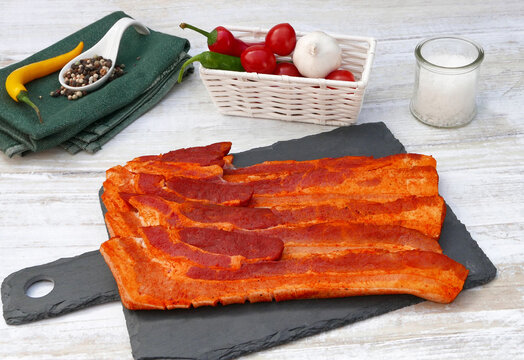 Rohe eingelegte Schweinebauchscheiben auf einer dunklen Schieferplatte. Im Hintergrund stehen ein K&ouml;rbchen mit Knoblauch, Tomaten und Peperoni sowie ein Salzstreuer auf einem hellen Holztisch.