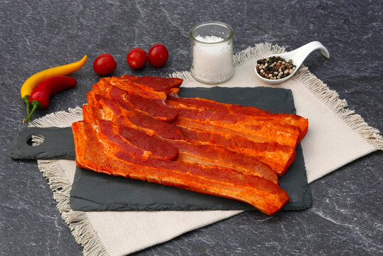 Rohe Schweinebauchscheiben auf einer dunklen Schieferplatte dekoriert mit Salzstreuer, Pfefferk&ouml;rnern  Kirschtomaten und Chilischoten auf einem dunklen Steinhintergrund.