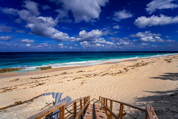 Fototapeta premium Wooden staircase leading down to a pristine sandy beach on Great Guana Cay, Bahamas, with turquoise Caribbean waters, gentle surf, and dramatic cloud formations under a vibrant blue tropical sky.