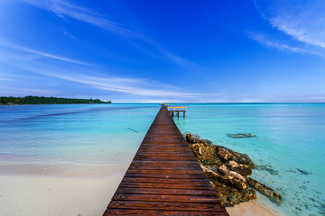 Obraz premium Long wooden pier stretching into crystal-clear turquoise waters in Cherokee, Abaco, Bahamas. Serene tropical seascape with pristine white sand beach, calm Caribbean lagoon, and dramatic blue sky.