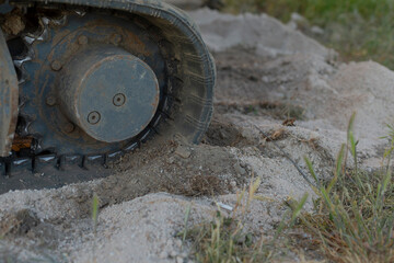 Close-up view of a large tracked vehicle wheel moving over sandy ground with dirt, gravel, and sparse grass in an outdoor setting, showing rugged equipment © Viktar