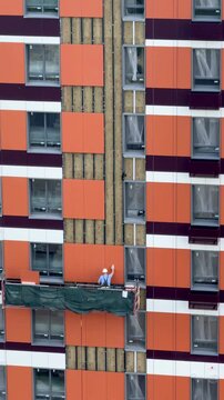Construction worker in hard hat installing vinyl siding on high-rise building facade from aerial lift, summer exterior finishing with safety harness