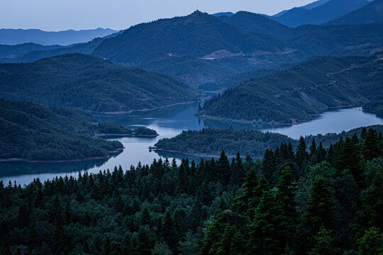 Panoramic twilight view of Lake Plastira in Greece, featuring winding blue waters surrounded by lush green pine forests and rolling mountain peaks under a soft, dusky evening sky.