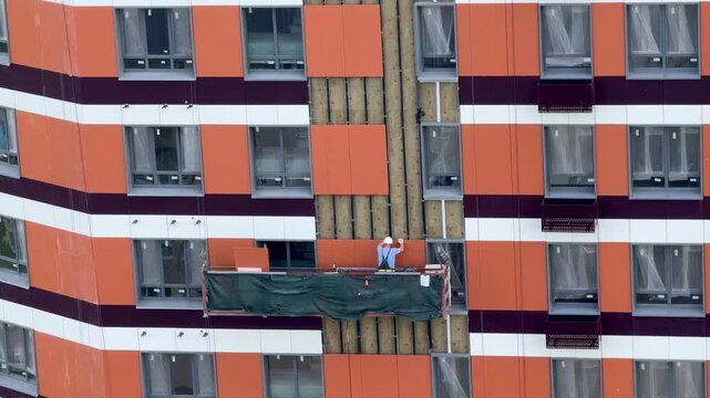 Construction worker in hard hat installing vinyl siding on high-rise building facade from aerial lift, summer exterior finishing with safety harness