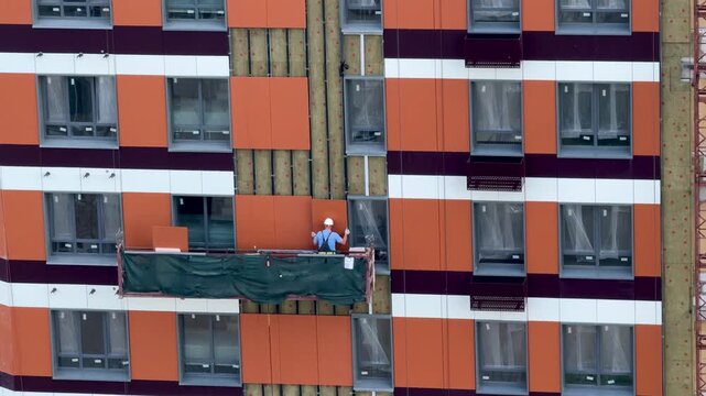 Construction worker in hard hat installing vinyl siding on high-rise building facade from aerial lift, summer exterior finishing with safety harness