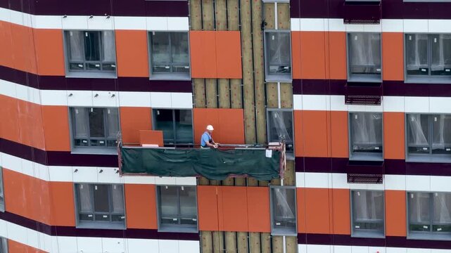 Construction worker in hard hat installing vinyl siding on high-rise building facade from aerial lift, summer exterior finishing with safety harness