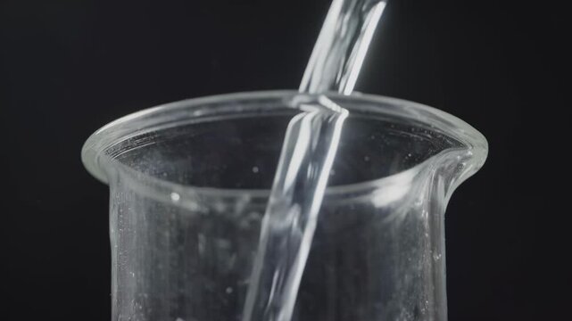 Transparent glass beaker positioned on a dark surface as clear water is poured in, showcasing the fluid's movement and interaction with the container's interior