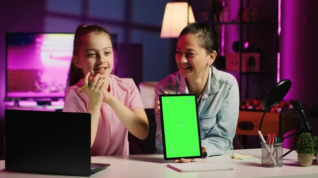Cute daughter filming internet show helped by her parent, promoting green screen tablet from sponsoring brand. Small child and her mom do influencer marketing, urging fans to purchase mockup device