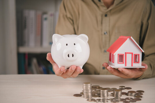 Man holding piggy bank and house model with stacked coins on table representing real estate investment property loan housing mortgage business finance wealth savings buying home concept