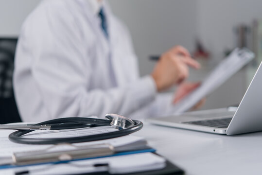 Stethoscope and clipboard on hospital desk with blurred male doctor reviewing medical records in background concept for health insurance healthcare administration and medical examination services