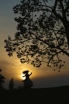 A silhouetted woman twirls and dances beneath a sprawling tree at sunset, framed against a warm golden sky that evokes calm, freedom, and evening serenity in a natural landscape...