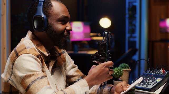 African american man interviewing celebrity, reviewing notes to ask pertinent questions. Journalist doing radio interview in apartment studio, wearing headphones and talking on mic with guest