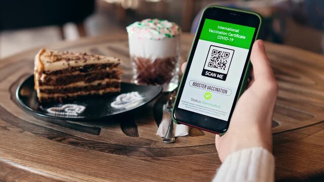 Person holding smartphone displaying international vaccination certificate with qr code in cafe.