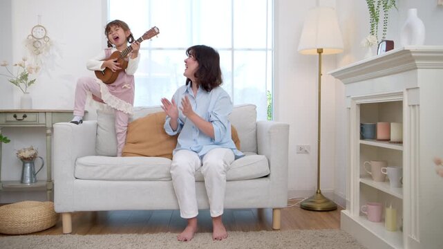 Little girl confidently playing the ukulele while her mother applauds