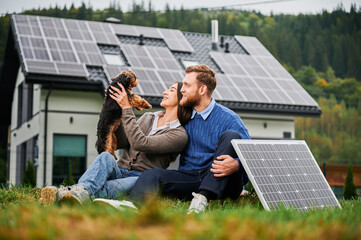 Couple sitting on grass playing with small dog near modern house with solar panels. Photovoltaic system on rooftop, renewable energy concept, eco friendly lifestyle © anatoliy_gleb