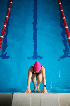 Young woman swimmer preparing for start freestyle backstroke in pool holding edge with both hands, looking down, wearing swimsuit, cap and goggles, indoor aquatic fitness concept