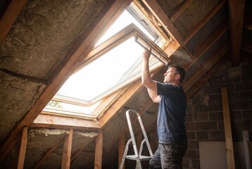 Fototapeta premium A man installing a skylight window in an attic, surrounded by insulation and wooden beams, bringing natural light into the space.
