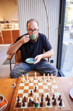 Man studying chessboard while sipping tea, intense focus on midgame position, upright posture and analytical gaze, coffee shop interior with muted tones, strategy practice and quiet concentration