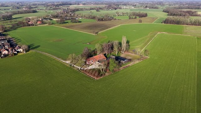 An aerial drone view showing a traditional farmstead with red-roofed buildings surrounded by green trees and sprawling patchwork agricultural fields under a clear sky.