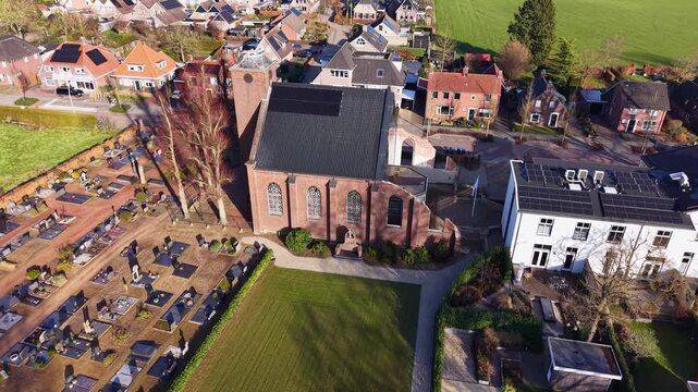 An aerial drone perspective of a historic brick church with an adjacent cemetery, showing residential houses with solar panels and a small village street under bright afternoon sunlight.