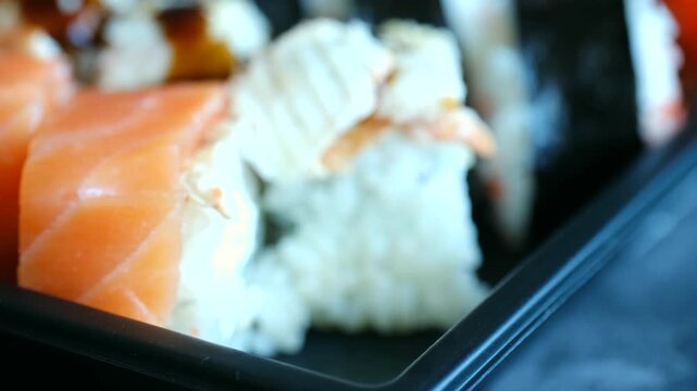 Close up view person taking fresh Philadelphia sushi roll piece from black plastic container with bare fingers, no chopsticks. Japanese food takeout concept. Hand picking up sushi roll bare fingers