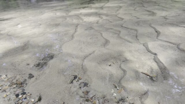 Clear water flows over a sandy riverbed forming small wavy ripples. Fine sand and tiny pebbles are visible through the transparent shallow stream in daylight.