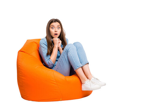 Full size photo of scared girl sit big lounge chair watch horror series feel nervous have shock stay speechless voiceless wear denim jeans outfit sneakers isolated white color background