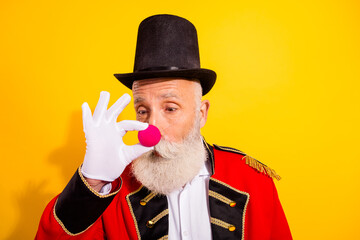 Elderly circus magician in a red coat and top hat performs a playful moment with a pink nose against a bright yellow background © deagreez