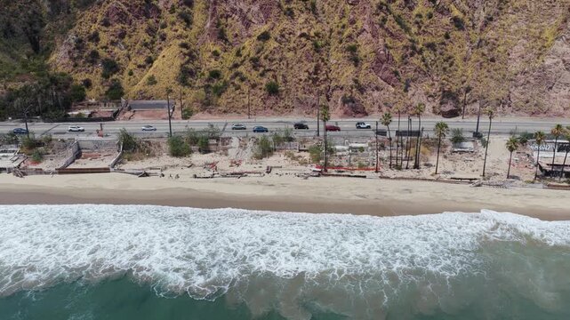 Cars driving on Pacific Coast Highway next to damaged Malibu real estate from Woolsey Fire and ocean waves crashing on sandy beach near Big Rock with palm trees