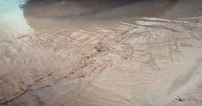 Slow motion close up: muddy runoff from a construction site flows rapidly, creating ripples while reflecting passing vehicles.