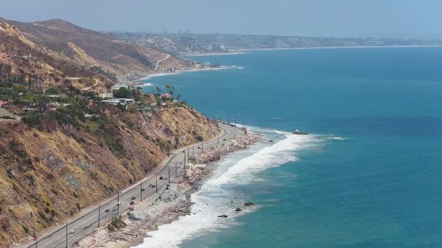 Malibu coastline showcasing Pacific Coast Highway with moving cars, oceanfront real estate, white waves breaking on shore under clear daytime conditions in California