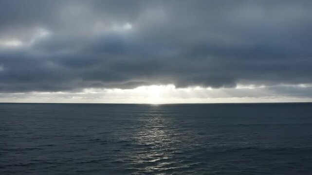 Sunlight breaks through dramatic clouds over a vast, calm ocean horizon.