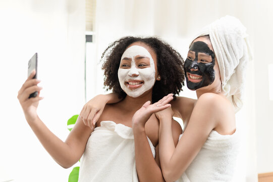 Young Asian and African woman using smartphone taking selfie photo together after applied beauty facial face mask at home. Young woman friendship daily lifestyle or LGBTQ couple concept.