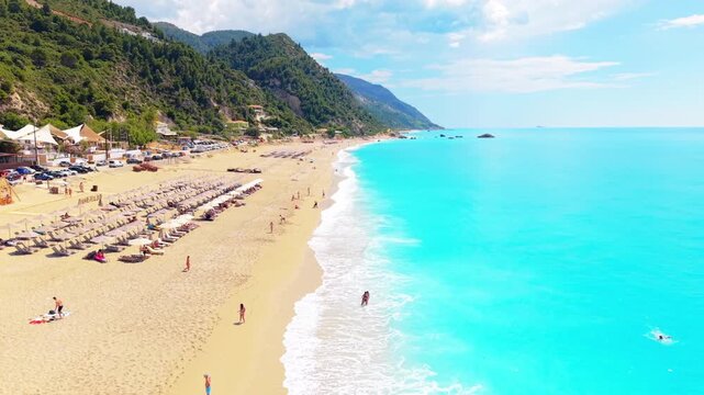 Drone flying forward along a stunning sandy beach with crystal-clear turquoise water and visitors enjoying a relaxing summer day by the sea. 