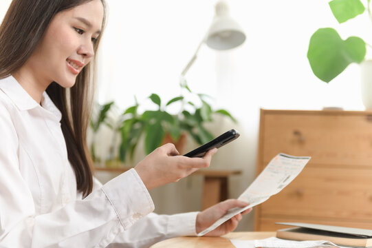 Young Asian woman using smartphone to scan a receipt for electronic payment and digital banking. Mobile finance, cashless transaction, fintech technology, online shopping and expense management.